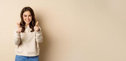Smiling happy woman choosing you, pointing fingers at camera and laughing, inviting people, standing over beige background photo