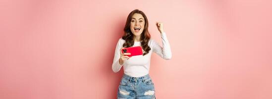 Beautiful girl looking enthusiastic, scream of joy and excitement, winning on mobile phone game, watching smth on smartphone and celebrating, pink background photo