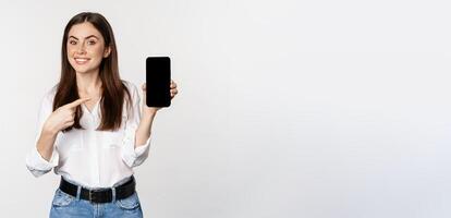 Happy beautiful woman smiling, pointing at mobile phone screen, showing advertisement, website, standing over white background photo