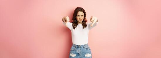 Portrait of young woman showing thumbs down to express dislike, disappointed, standing over pink background photo