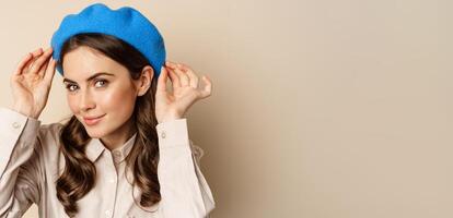 Stylish modern girl put on trendy hat on head and smiling, going out, posing against beige background photo