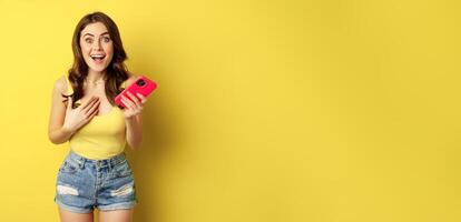 Surprised smiling woman holding mobile phone, reacting amazed and impressed at camera, seeing something awesome, standing against yellow background photo