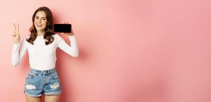 Stylish young woman introducing company website, mobile app, showing smartphone horizontally and peace sign, smiling happy, standing over pink background photo