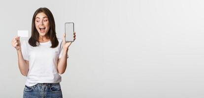 Excited attractive girl showing smartphone screen and credit card, white background photo