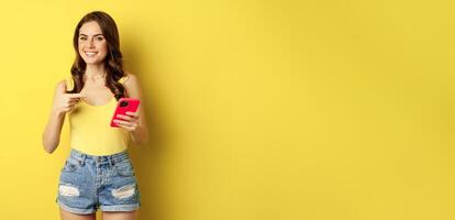 Cellular technology and online shopping concept. Beautiful smiling woman pointing finger at her mobile phone, recommending website, app on smartphone photo