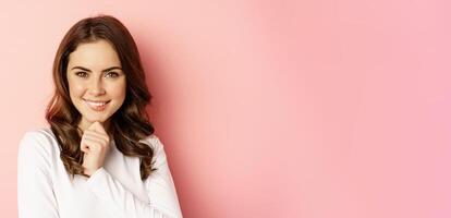 Close up portrait of thinking girl, smiling and looking confident, thoughtful face expression, standing over pink background photo