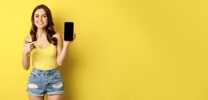 Young stylish woman showing mobile phone screen, pointing at smartphone display, app interface or online store, smiling pleased, standing over yellow background photo