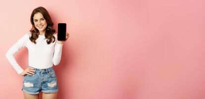 Stylish sexy woman showing mobile phone screen, smartphone app interface and smiling, demonstarting online shop, standing over pink background photo