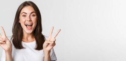Portrait of beautiful and happy brunette girl smiling and showing peace gestures, standing white background excited photo