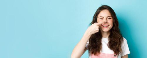 Close up of funny brunette girl touching her nose and smiling cheerfully at camera, standing against blue background photo