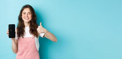 This is good. Smiling cheerful woman showng empty smartphone screen and thumb up to recommend app or online store, standing against blue background photo