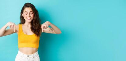 Portrait of stylish natural girl in summer crop top, pointing and looking down with dreamy smile, seeing something interesting on bottom advertisement, blue background photo