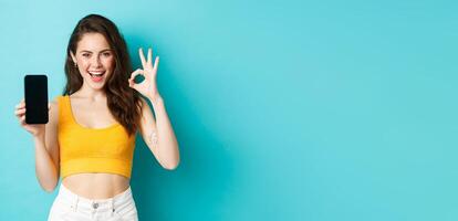 Look at this. Good-looking woman with sassy smile, winking and showing okay sign with empty smartphone screen, recommending app, standing over blue background photo
