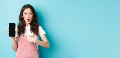 Impressed young modern woman pointing at smartphone screen, say wow and stare amazed at camera, showing awesome application, new online store, standing over blue background photo