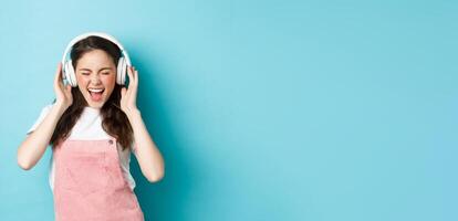 Carefree pretty girl scream from joy, listening music in headphones with happy face, lip-sync lyrics, standing over blue background photo