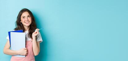 Beautiful girl student looking thoughtful at top copy space, standing with notebooks homework and smiling pensive, standing against blue background photo