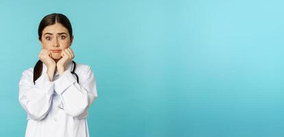 Scared and worried woman doctor in white coat, looking anxious and insecure, shaking from fear, standing over torquoise background photo