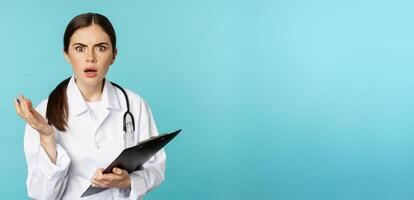 Shocked woman doctor reacting worried and frustrated at camera, hear smth strange, standing in white coat against torquoise background photo
