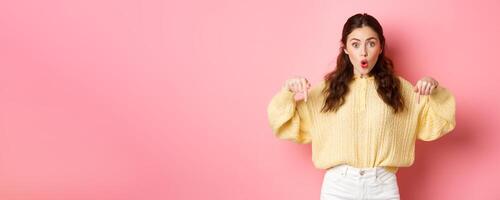 Wow super cool. Excited brunette girl pointing fingers down, gasping wondered, showing direction way, indicating at bottom logo, standing over pink background photo