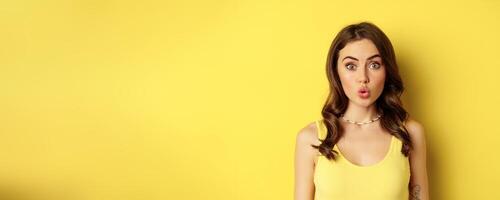 Close up portrait of stylish brunette girl looking amazed, wow face, showing excitement and surprise, impressed by something, standing over yellow background photo