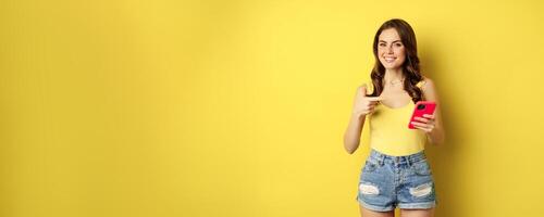 Cellular technology and online shopping concept. Beautiful smiling woman pointing finger at her mobile phone, recommending website, app on smartphone photo