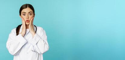 Portrait of shocked female doctor, worried healtcare worker in white robe, looking concerned at patient, standing over blue background photo