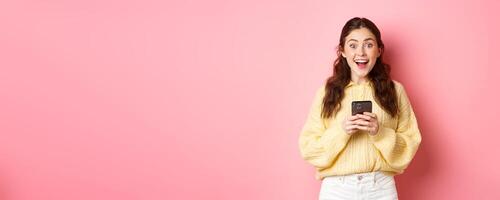 Portrait of girl holding smartphone, looking surprised and gasping excited, found something interesting online, standing against pink background photo