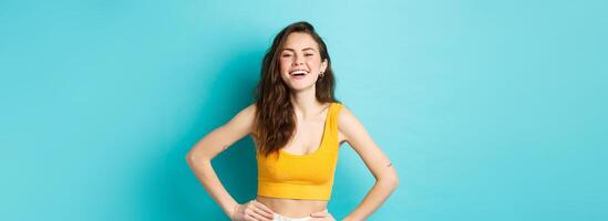 Image of confident and happy stylish woman laughing, holding hands on waist and smiling cheerful, standing carefree in summer outfit, blue background photo