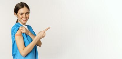 Smiling nurse, female healthcare worker in scrubs, pointing finger right, showing patch on shoulder, vaccination campaign, covid-19 and pandemic concept, white background photo