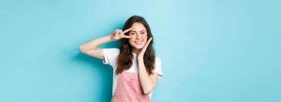 Portrait of kawaii teenage girl giggle and showing peace v-sign near eye, smiling cheerful and touching cheek, standing over blue background photo