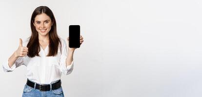 Smiling woman showing thumbs up, recommending app, mobile phone screen, empty interface on smartphone, standing against whte background photo