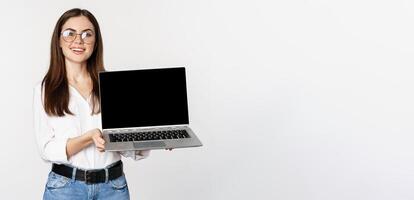 Young woman in glasses showing laptop screen, demonstrating promo on computer, website or store, standing over white background photo