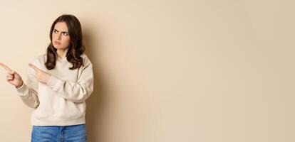 Woman pointing left and staring confused at banner copy space, strange logo, standing over beige background photo
