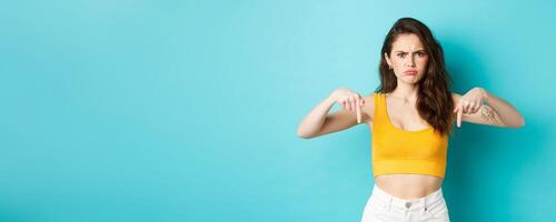 Confused frowning woman looking displeased, need explanation, pointing fingers down at something strange or unpleasant, standing over blue background photo