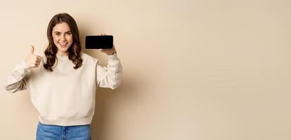 Smiling happy woman showing smartphone horizontal screen, thumbs up, recommending website, online store or app, standing over beige background photo