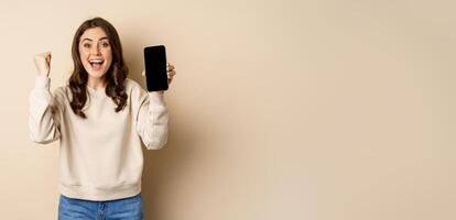 Enthusiastic woman cheering and showing cellphone screen, smartphone app interface, standing over beige background photo