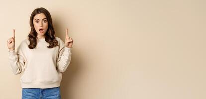 Surprised beautiful girl showing advertisement, pointing fingers on top, store sale or website link up, standing over beige background photo