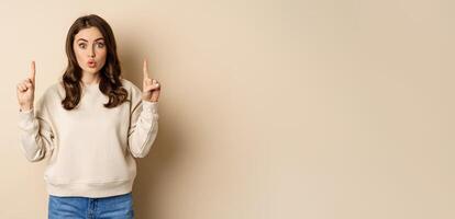 Surprised beautiful girl showing advertisement, pointing fingers on top, store sale or website link up, standing over beige background photo