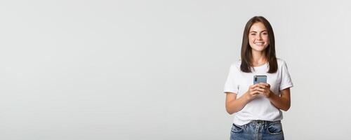 Attractive smiling brunette girl texting message, using mobile phone and looking at camera photo