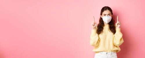 Covid, corona and social distancing concept. Confused and shocked young woman in medical respirator pointing fingers up, staring startled at camera, pink background photo