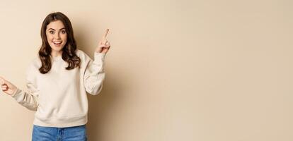 Cheerful caucasian woman showing directions, two ways, pointing sideways at variants, choices for customer, standing over beige background photo