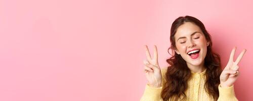 Positive emotions. Beautiful young woman showing peace v-sign and smiling carefree, posing with eyes closed and happy face, standing against pink background photo