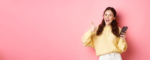 Portrait of attractive glamour girl showing thumbs up after using app on smartphone, online shopping application, standing against pink background, smiling satisfied photo