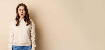 Portrait of shocked brunette woman drop jaw, gasping and staring speechless at camera, beige background photo