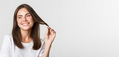 Portrait of flirty beautiful girl playing with hair and smiling at camera coquettish, standing white background photo