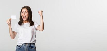 Joyful good-looking girl rejoicing and looking at credit card, fist pump while triumphing, white background photo