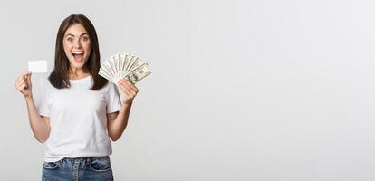 Excited happy brunette girl showing credit card and money, smiling over white background photo