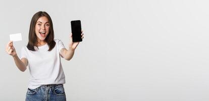 Attractive smiling girl looking satisfied and showing credit card, mobile phone screen photo