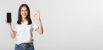 Pleased smiling brunette girl showing smartphone screen and showing okay gesture in approval photo