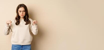 Sad and disappointed girl pointing, looking down with upset grimace moody face, standing over beige background photo